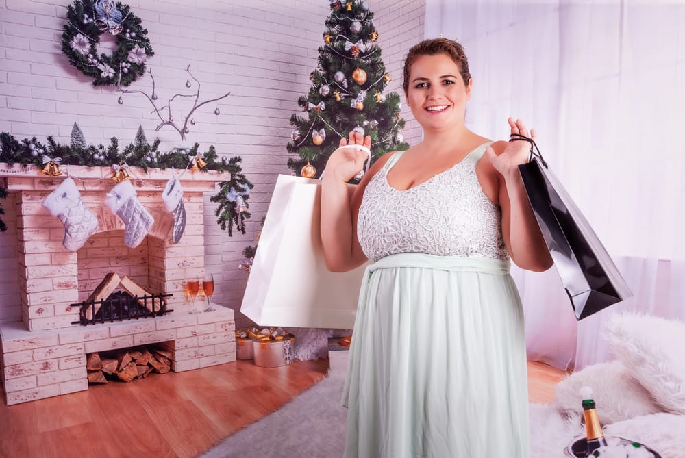 plus size woman in eloquent dress holding christmas gift bags with tree and fireplace behind her