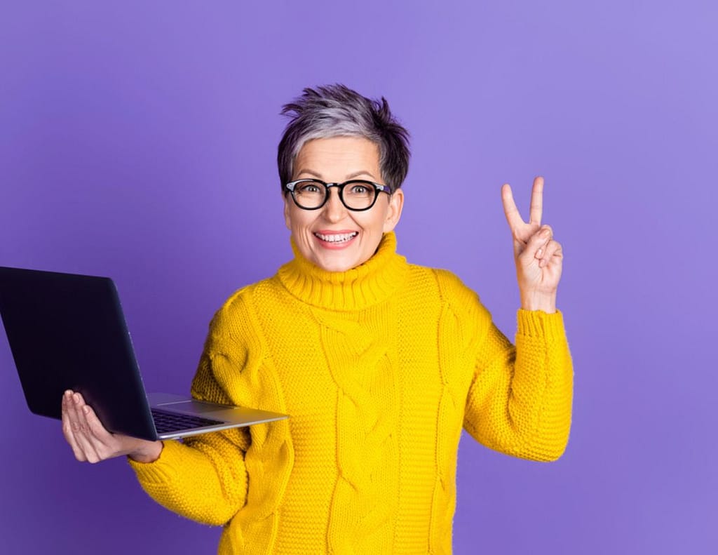 midlife woman yellow sweater laptop in hand purple backdrop